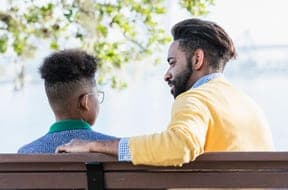 A man talking with a teenage boy on a park bench by the water