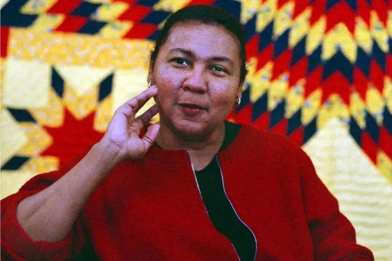 bell hooks in a red jacket, one hand raised to her cheek, in front of a star quilt