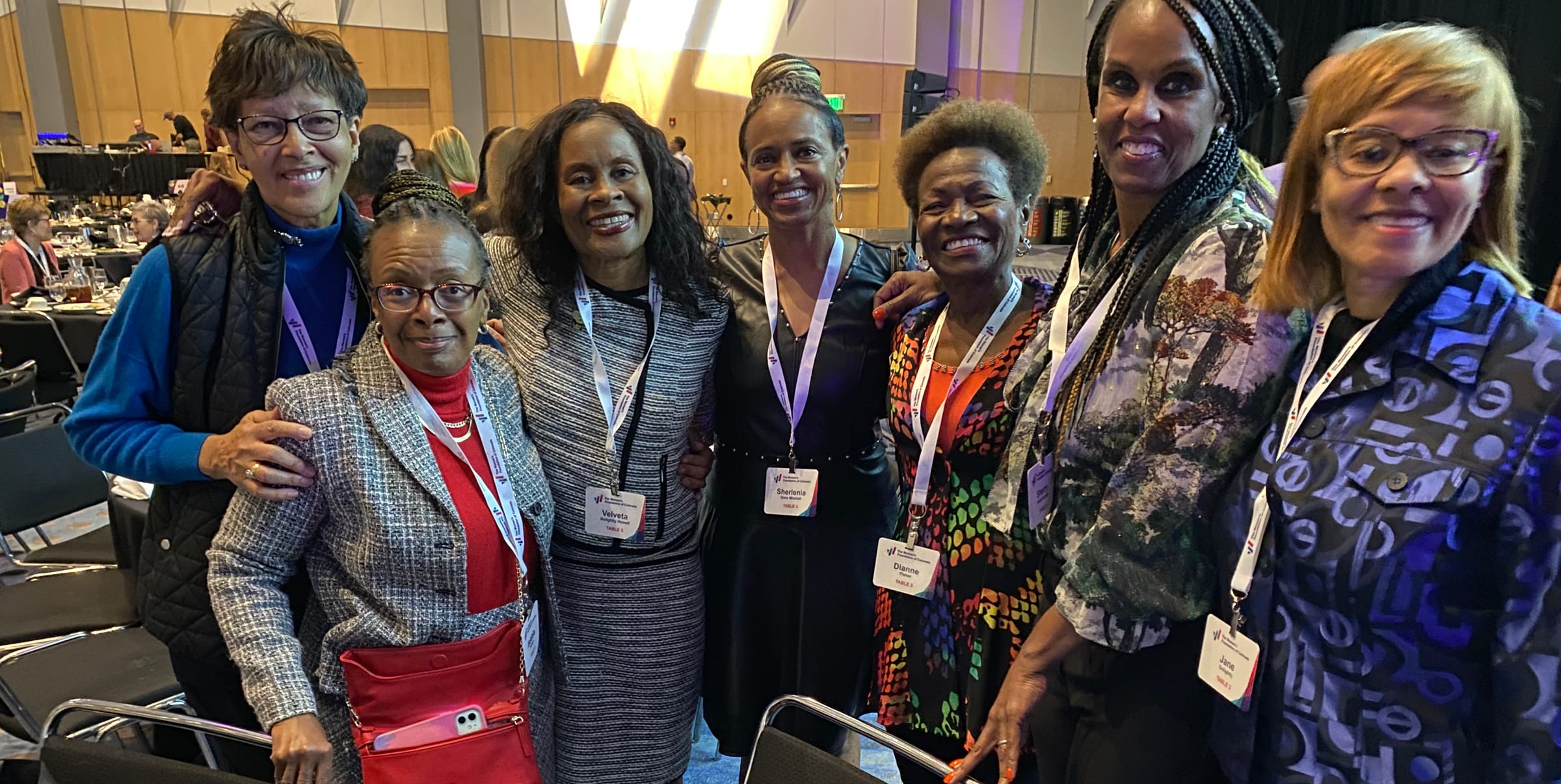 Seven Sister-to-Sister members with event lanyards, smiling together at a luncheon