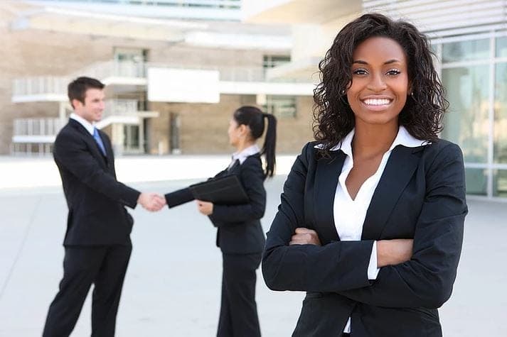 A smiling Black businesswoman in a suit, arms crossed, outside an office building while two colleagues shake hands behind her
